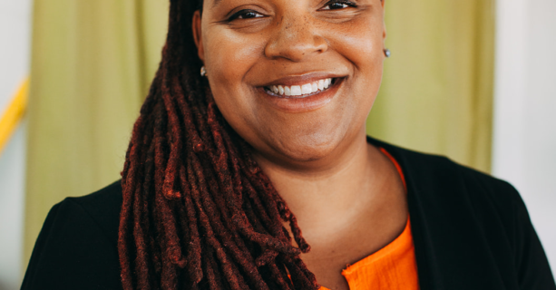 Kandace smiles at the camera with a yellow background behind her. She is a Black woman with long locs. She wears a black blazer with an orange blouse underneath.