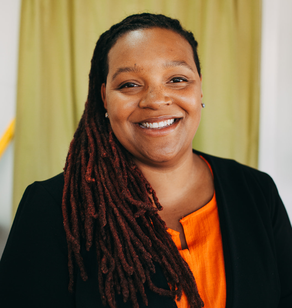 Kandace smiles at the camera with a yellow background behind her. She is a Black woman with long locs. She wears a black blazer with an orange blouse underneath. 
